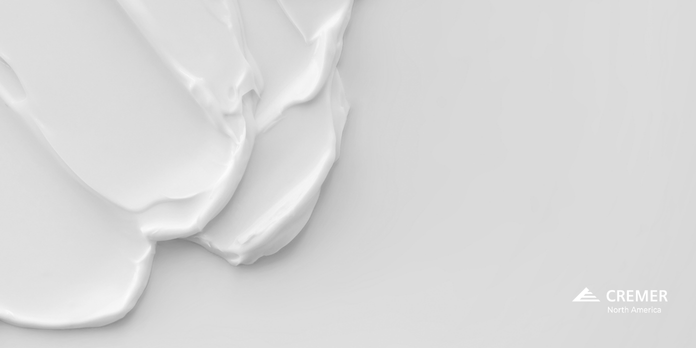 Close-up of a smooth, glossy white cream spread across a light gray surface, showing its thick texture; a small ‘CREMER North America’ logo appears in the bottom right corner.